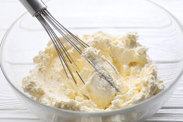 Bowl with whisk and whipped cream on white wooden table, closeup