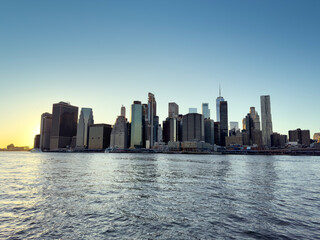 Obraz premium Panoramic View of Lower Manhattan Skyline at Sunset with Reflections on the East River and Clear Evening Sky – Iconic Urban Silhouette of New York City