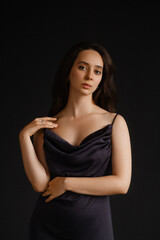 Caucasian woman in black satin dress on dark background. Touching shoulder, front view, looking into camera. Concepts of beauty, skincare, cosmetics, self-care, and luxury fashion.