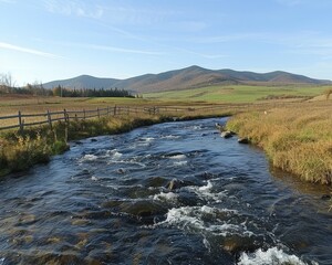 Fototapeta premium Scenic river flows past meadows and mountains under a blue sky, bordered by a rustic wooden fence
