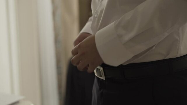 A young male businessman fastens a belt on his trousers while standing at the window going to a business conference
