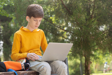 A teenage boy in a yellow hoodie sits in a park with a laptop, wearing wireless headphones. Green...