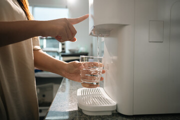Woman using a modern water purifier in kitchen, filling a glass with fresh, purified water