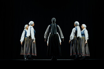 A group of dancers in traditional attire stands in formation on a dark stage, showcasing cultural...