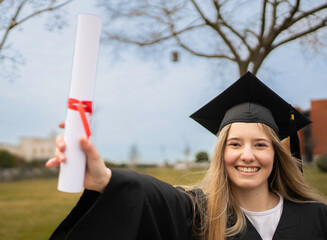 Portrait of smiling graduate showing diploma tied with red ribbon, wearing graduation gown and cap,...