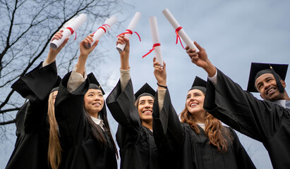 Happy multiethnic graduates raising diplomas outdoors wearing cap and gown against blue sky