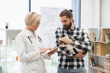 Female veterinarian discussing treatment plan with adult bearded man holding cat in clinic. Medical professional explaining pet care using tablet demonstrating attentive and professional consultation © sofiko14