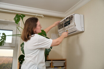 Woman repairing wall-mounted air conditioner with screwdriver in a home interior. HVAC maintenance...