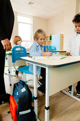 A blonde White school-aged boy wearing a blue shirt studies at his desk with a Black classmate and female teacher standing to his sides, in a classroom with beige walls and a window in the background.