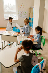 Obraz premium Diverse elementary school students work on tasks like drawing in a classroom. The room features wooden desks, colorful chairs, and educational materials in a room with white walls and a window.