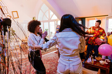 Diverse friends celebrate a birthday party in a room with large windows and white walls. There are drinks and balloons in the room. One of them holds a cake with sparklers near a wooden table.