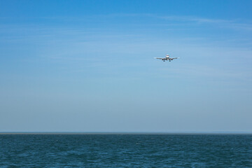 An airplane comes in for a landing over the sea under a clear blue sky. The aircraft is flying low with its landing gear released, preparing to touch down