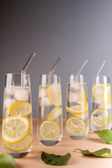 four tall glasses filled with refreshing lemon-lime water adorned with ice cubes and glass straws, positioned on a beige surface against a gradient blue background with scattered green leaves.