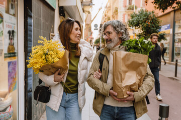 A middle-aged European couple in their late 50s, walking together through a city street and talking, as the husband holds a paper bag of groceries and the wife holds a bouquet of simple flowers
