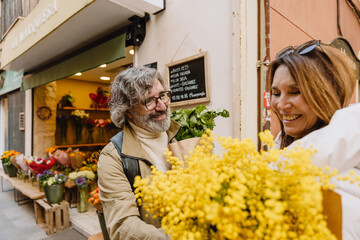 A middle-aged White husband in his late 50s, smiling as he gifts a bouquet of simple yellow flowers to his middle-aged White wife, as they stand together on the city street near a flower vendor