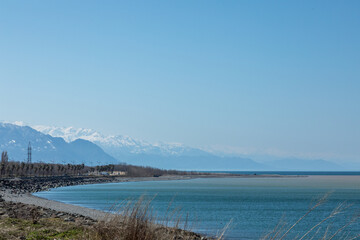 A picturesque coastal landscape with a pebble beach, calm blue sea, and distant snow-capped mountains under a clear sky
