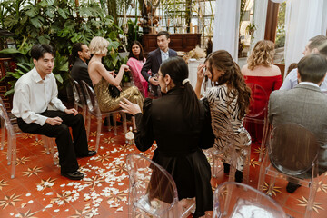 A diverse group of elegantly dressed guests sit on clear chairs at a wedding reception, surrounded by greenery and petals, engaging in conversation. An adult Asian man talks with an Asian woman.