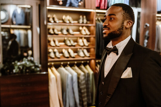 A Black man in his 30s with short black hair and a beard dressed in a white shirt, a bow, and a black blazer standing in a shop with blazers and shoes in the background during try-on a groom's suit