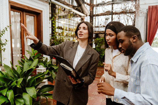 A White female location scout in her 30s, talking to a young Black couple who are in their 20s, and gesturing around as they stand inside of a spacious room with various potted plants around them