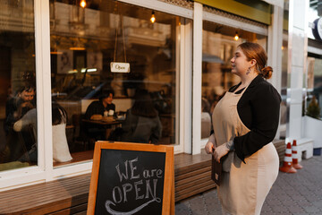 A young, overweight White woman in her late 20s, who is wearing a beige apron, standing outside of a small coffee shop, near a chalkboard sign, and looking at the building