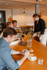 An adult White woman with red hair wearing a black blouse and beige pants is talking to three adult White men in a cafe, one in a striped sweater, others using laptops and phones, all smiling.