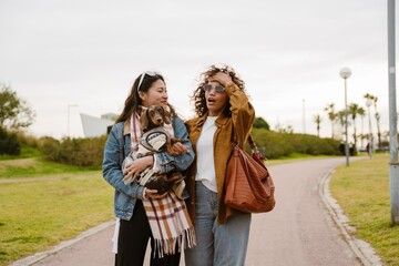 Fototapeta premium A young Latina woman in her late 20s, smacking her forehead in astonishment as she walks together with her young Asian female friend on a walkway in a grassy park, her friend carrying a dachshund dog