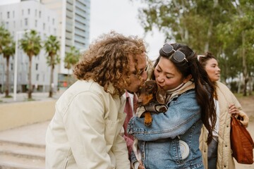 Obraz premium An adult Asian woman and an adult White man wearing sunglasses hold a dachshund on a street with buildings in the background. They are kissing the dog. An adult Latino woman stands in the background.