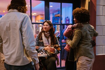 A group of multiethnic friends, with an adult Asian woman with a beige scarf holding a dachshund, gathers in front of a cafe featuring colorful lighting and a window. They are talking and hugging.