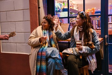 An adult Latino woman and an Asian woman in her 30s sit outside a bar with drinks and a dog, laughing and interacting with a Black man on the left. The cafe facade has white walls.