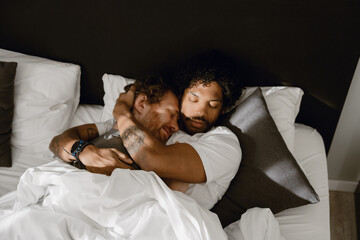 In a bedroom, a gay couple in their 20s, a smiling White brown-haired man and a curly Black brunet...