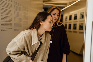 Two adult friends walking in the museum with photos on the walls, a woman with brown hair in a...