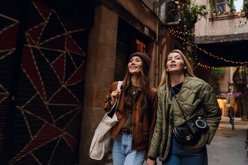 A young White woman in her late 20s, with a camera around her neck, holding her young White girlfriend by the hand and looking around as they walk along a beautiful alleyway outside, on a sunny day