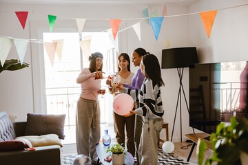 A group of four women in their late 20s dressed in bright sweaters and jeans standing with wine glasses in a bright room with a colorful garland, a woman on the left pouring wine from a bottle
