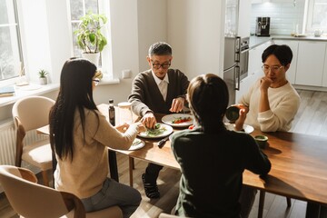 An Asian family gathers around a table. They are eating a meal together in a bright, inviting kitchen space with white windows. An elderly Asian grandfather takes vegetables from a plate.
