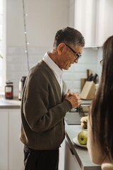 Fototapeta premium An elderly Asian grandfather in glasses and a sweater stands in a kitchen, with a plate of fruit on the counter, and smiles. His adult daughter talks to him. The kitchen has white walls and counters.