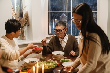 An elderly Asian grandpa in his late 70s, holding chopsticks and talking to his mid-20s grandson, as they sit at the dinner table together, eating, as his middle-aged daughter sets more plates