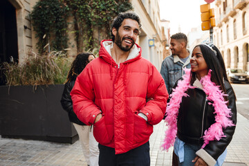 Naklejka premium A Latino man in his late 20s with short black hair and a beard in a red jacket walking on the street with a Black young adult woman in a crown and a pink feather scarf, and two adult Latino friends