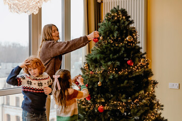 A White mother in her 30s, decorating a Christmas tree and smiling as her two school-aged kids, a boy, and a girl, help her pick up the decorations, in a cozy apartment near the windows