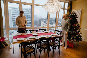 A middle-aged White son and an elderly Caucasian father stand in a festively decorated dining room with a Christmas tree, chandelier, and panoramic city view, captured through large windows.