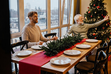 A White man in his late 30s, sitting at a long wooden table together with his elderly White father, as they're setting up for Christmas dinner, in a cozy apartment with a Christmas tree beside them