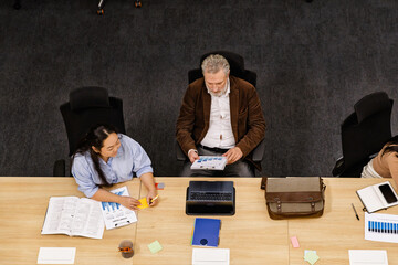 A calm elderly White man and an adult Asian woman review documents and use a laptop at a long wooden table in an office, surrounded by office supplies and a briefcase during a business meeting.