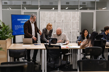 A White businesswoman in her 30s, helping her White male colleague fill out some paperwork while standing next to him as he sits among their other colleagues at a wooden desk, in an office room
