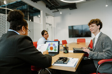 A young White man in his late 20s, smiling happily as he talks to his older Black female coworker while she sits at the head of a table in an office meeting room, as he sits beside her