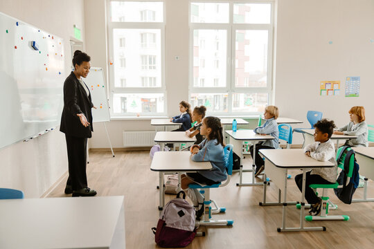 A Black female teacher in her 30s stands next to a whiteboard and talks to a multiethnic group of children sitting at their desks in a classroom with white walls and large windows in the background.