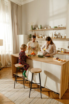 A White family prepares breakfast together in a kitchen. A school-aged son sits on a stool at a wooden counter, while the adult father and mother prepare food. Open shelves hold kitchen items.