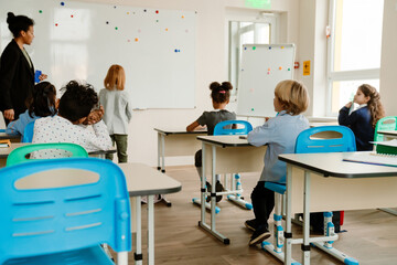 In a classroom with windows, a Black female teacher in her 30s stands near a whiteboard with one of her school-aged pupils who has medium-length red hair, with other children sitting at their desks.