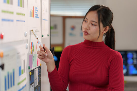 Asian businesswoman wearing a red turtleneck sweater is pointing at charts and graphs on a whiteboard, analyzing data and discussing business performance in an office meeting room