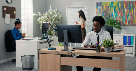 A man in a white shirt and tie works with focus at his computer, while in the background, his...