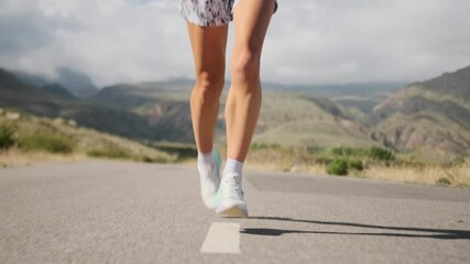 A woman in athletic wear and sunglasses runs along a scenic road, her muscular legs confidently covering the distance. Moving swiftly through majestic mountains, she building strength and endurance.