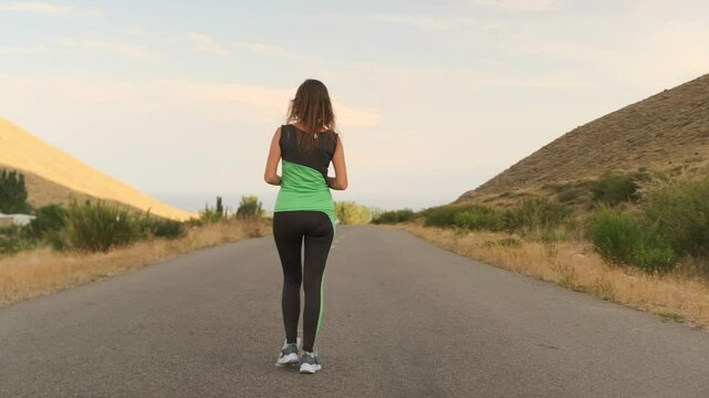 A young woman walks along a deserted road at the foot of the hills, selecting upbeat music for motivation. With a steady pace and cool evening breeze, she focuses and prepares for the run ahead.